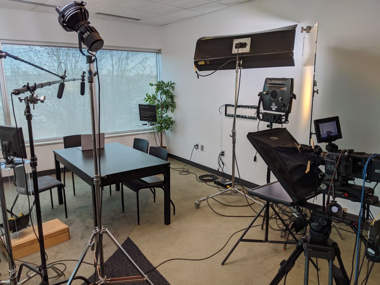 A room lit for an interview like a film set — overhead soft light, a hard fresnel key, teleprompter, and a cinema camera on sticks.