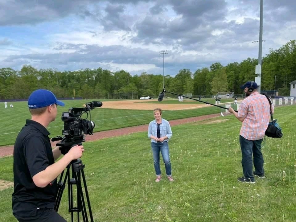 A three-person crew filming an on-camera interview on a baseball field — camera operator, subject, and boom operator.